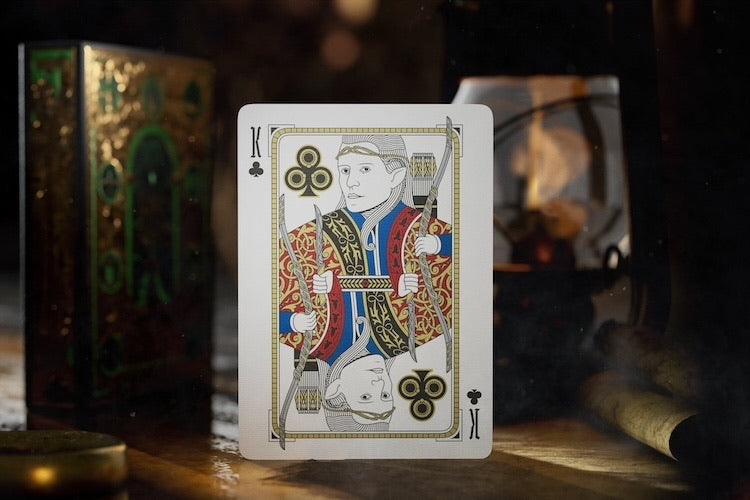 A single playing card stands upright on a wooden surface in a dimly lit, atmospheric setting. The card shows a stylized figure in ornate red, blue, and gold clothing with black clover motifs and the number 10 in the corners.
